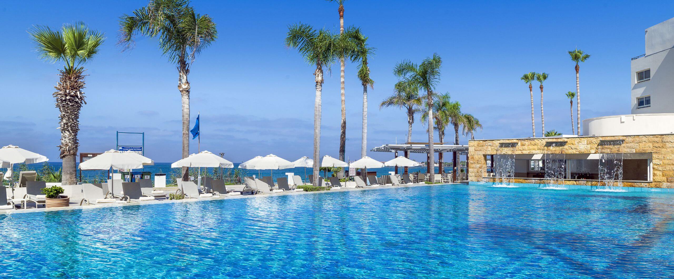 infinity outdoor pool area at Alexander The Great Beach Hotel, featuring cascading water jets, sun loungers with towels, palm trees, and panoramic views of the Mediterranean Sea