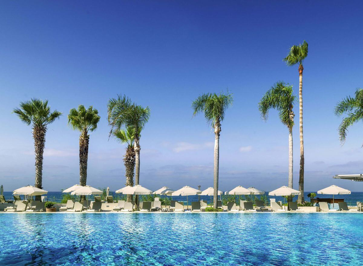 Tranquil outdoor pool surrounded by loungers and tropical greenery, with a clear view of the Mediterranean under a sunny blue sky.