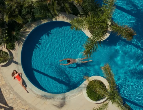 Leisure pool top-down view with a man swimming laps and a woman sunbathing on the stone deck surrounded by palm trees, Olympic Lagoon Resort Paphos