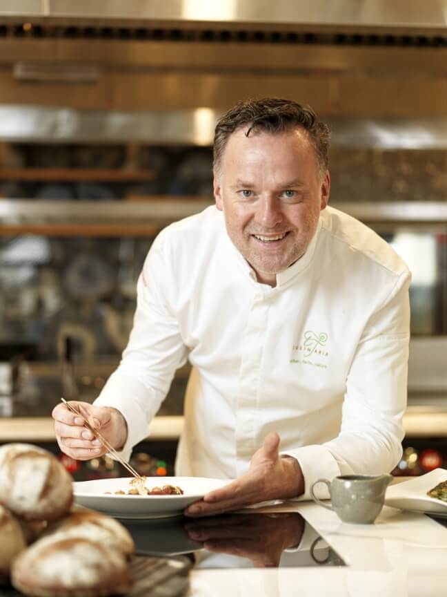 A chef in a white chef coat leaning over a pass counter in a kitchen, holding a pair of culinary tweezers while garnishing a plated dish next to freshly baked loaves of bread.